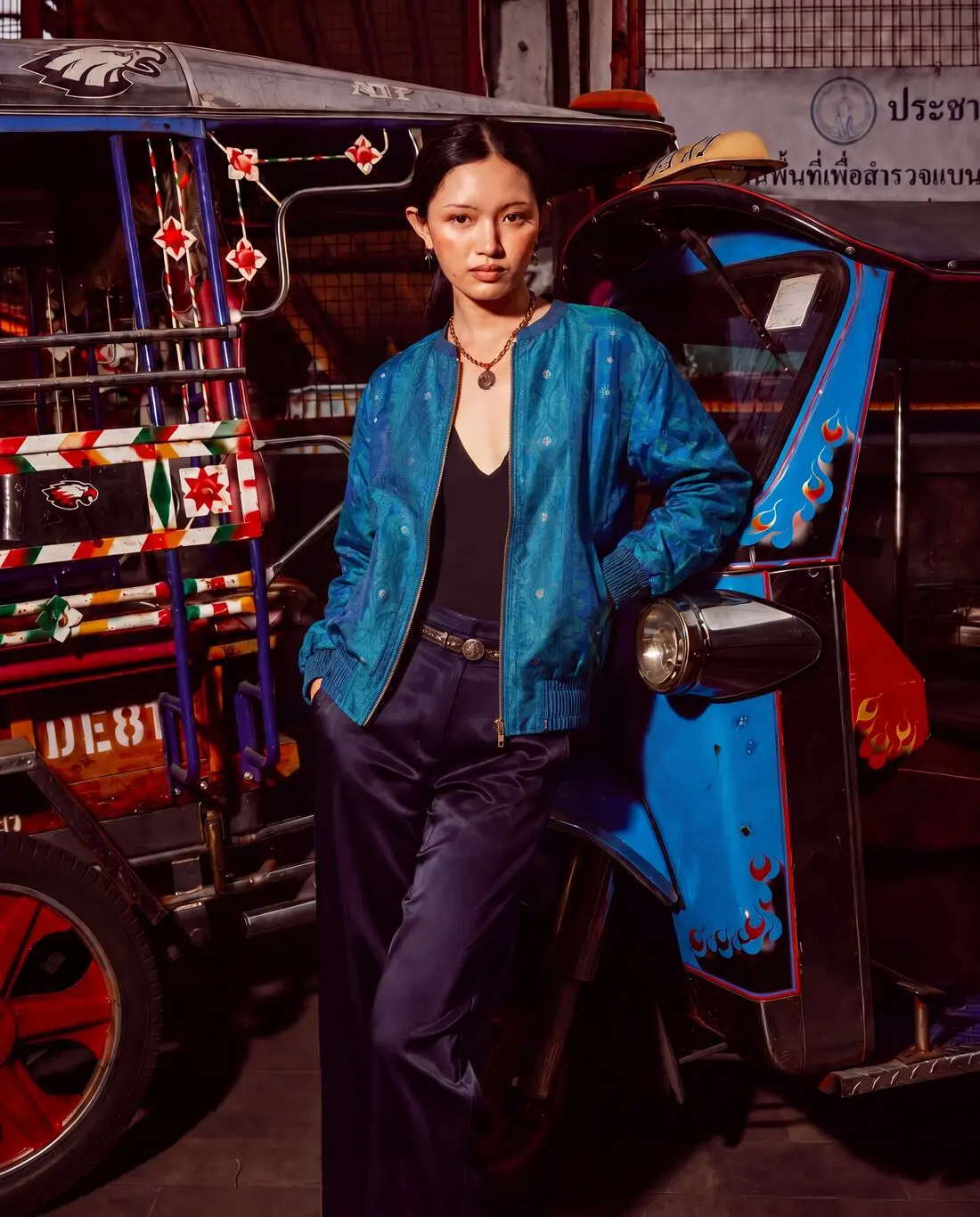 Model in a blue jacket leaning on a painted tuk-tuk at night